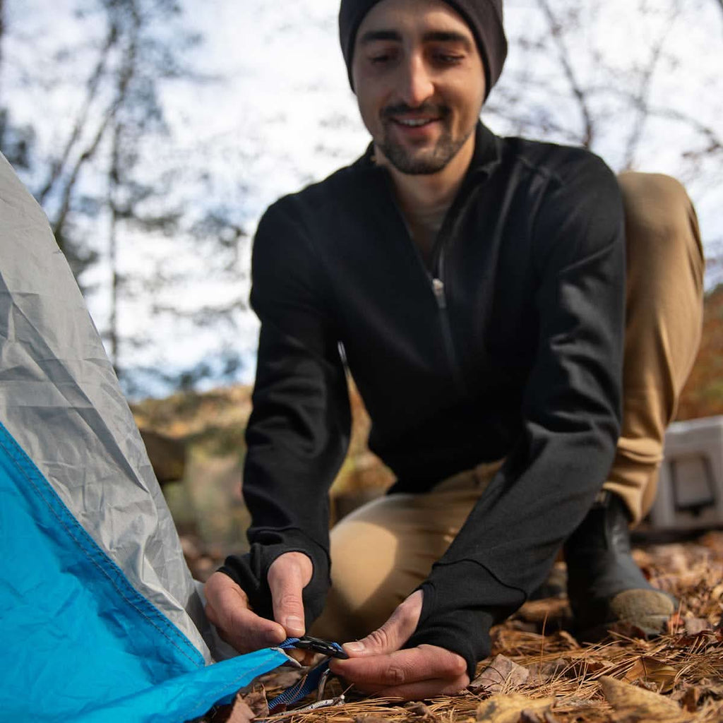 A man wearing a black 100 percent merino wool full zip sweater and beanie rigs up a tent in the woods