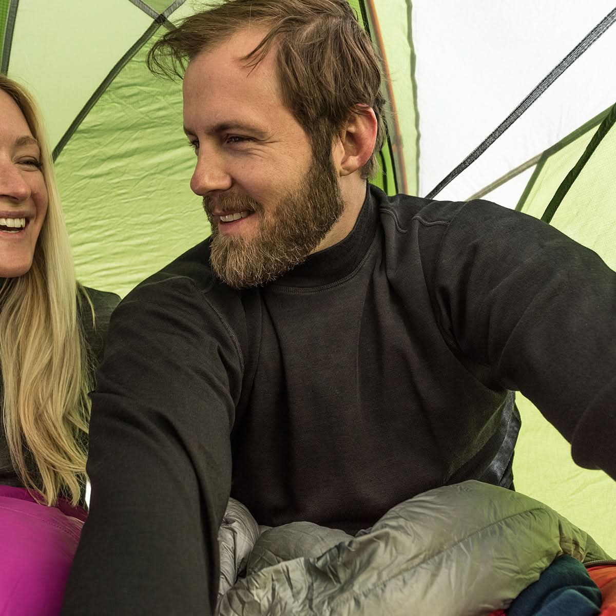 A bearded man wearing a black 100 percent merino wool mock neck smiling inside a green tent next to a woman.