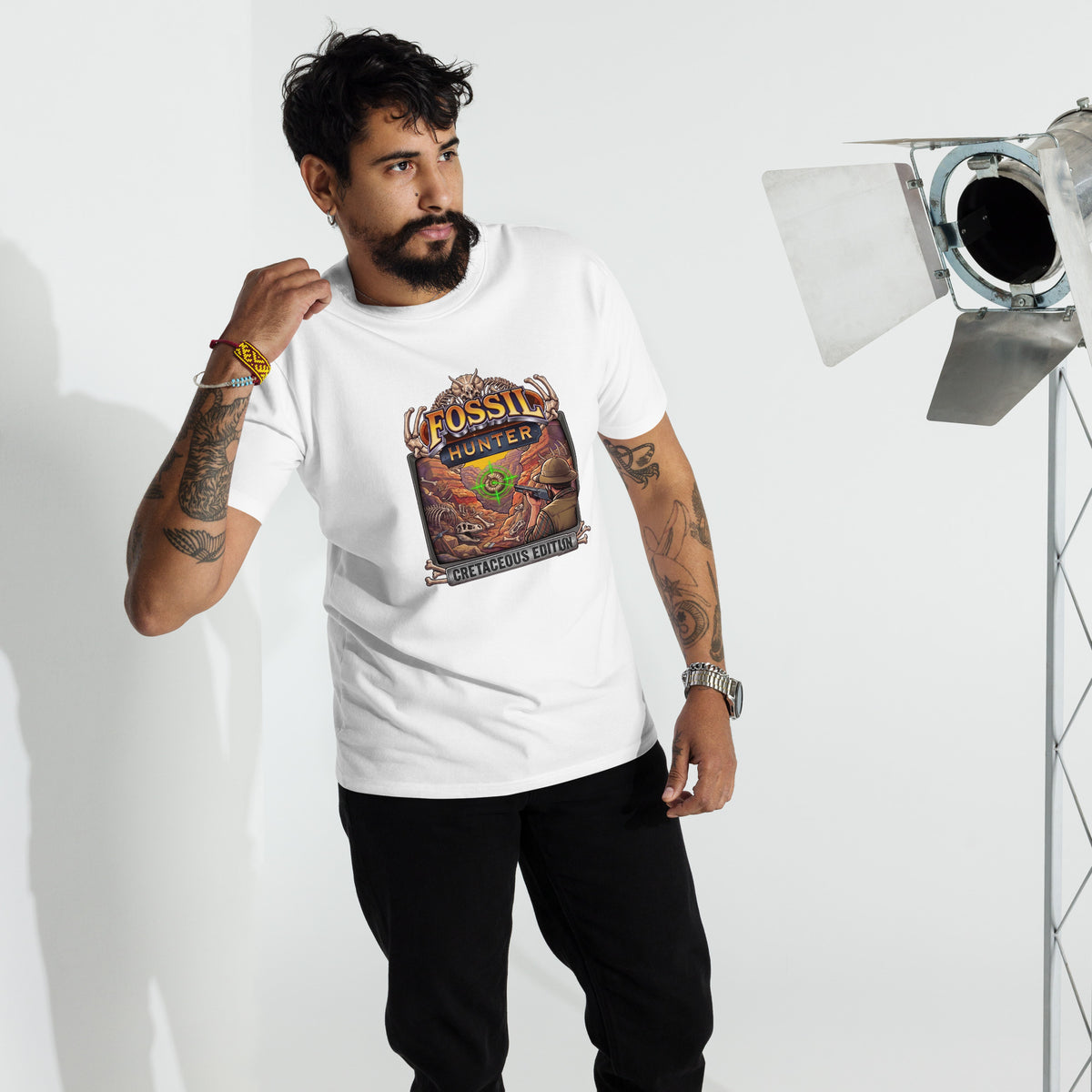 A bearded model wearing a white 100% cotton short-sleeve crewneck graphic t-shirt printed direct to garment on the front with a colorful graphic of a paleontologist fossil hunter targeting ammonite in his crosshairs with a pump action shotgun in a parody of classic arcade hunting games, standing in a studio setting with lights. 
