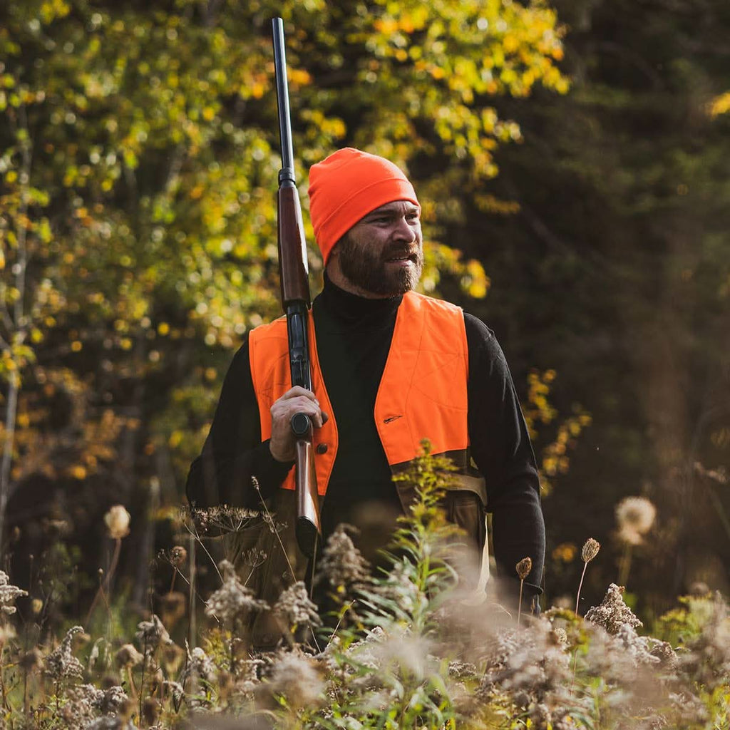 A bearded man wearing a black 100 percent merino wool turtleneck under a blaze orange hunting vest in the autumn woods.