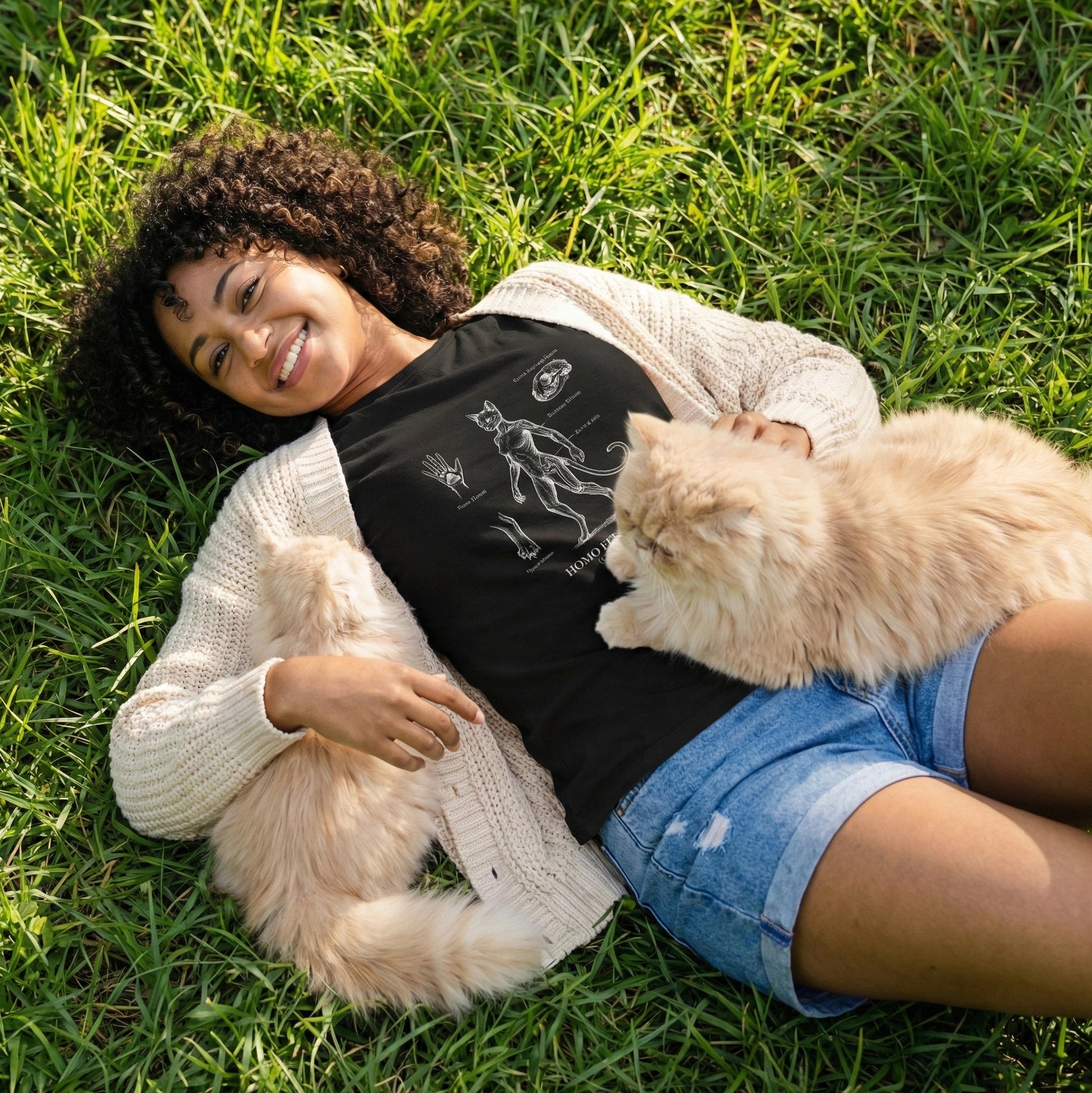 Woman lying on grass with a cat, wearing a black t-shirt and denim shorts.