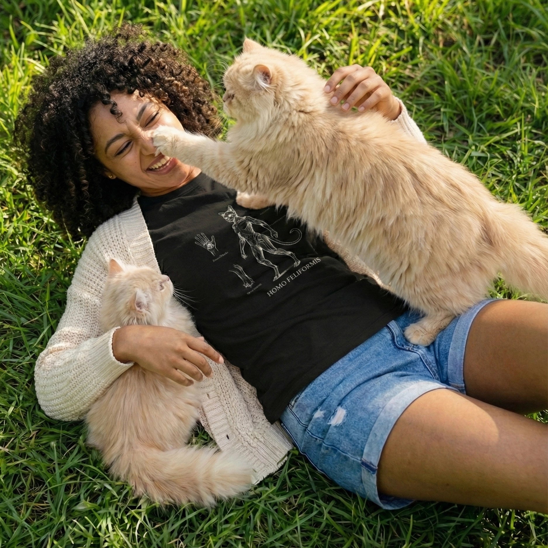 Woman lying on grass with two cats, one of which is playfully interacting with her face.