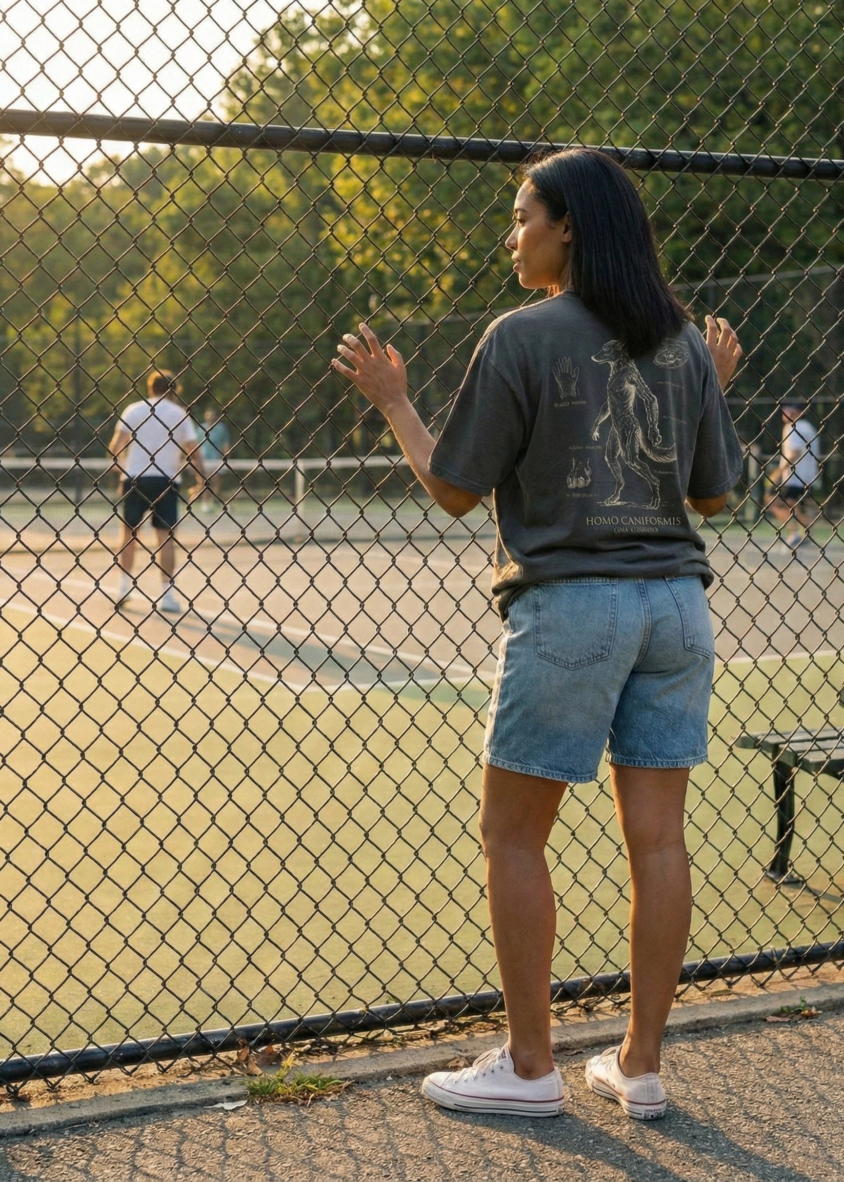 Person standing on a tennis court, looking over a fence at other players.