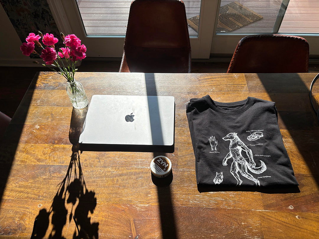 A black heavyweight all cotton graphic t-shirt with a white illustration of an antique anatomical drawing of a hybrid man-dog,next to a closed silver macbook, a vase of pink flowers, and a canister of nicotine pouches. The scene is shot from above in natural sunlight with strong shadows.
