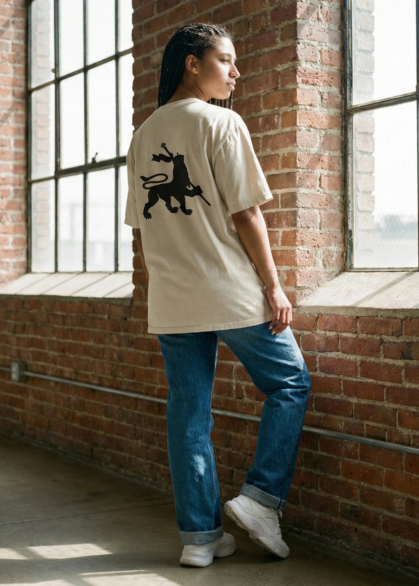 Person wearing a beige oversizedt-shirt with a black Lion of Judah rastafarian graphic design, standing against a brick wall.