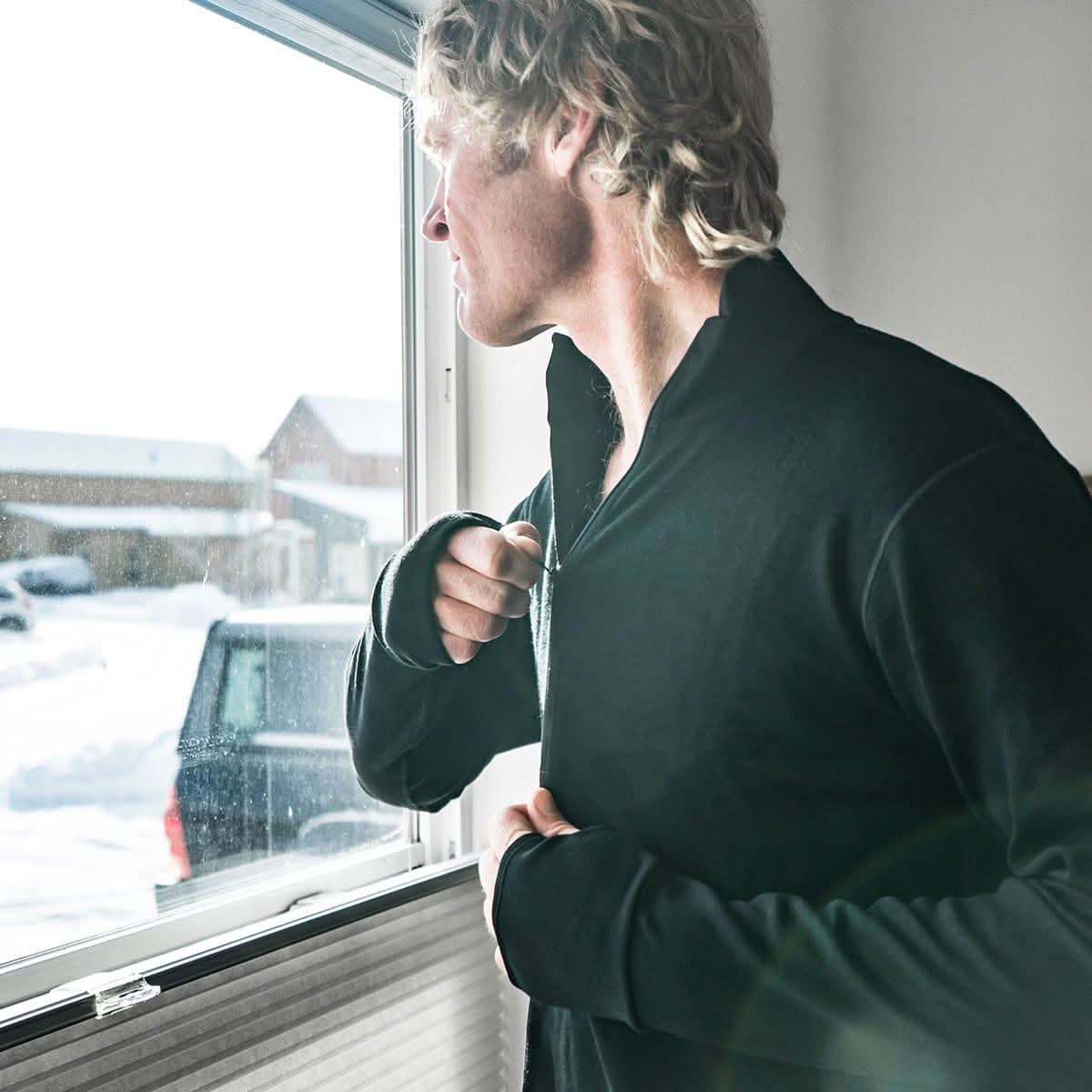 A man wearing a black 100 percent merino wool athletic quarter zip adjusting the collar while looking out a window at a snowy landscape.