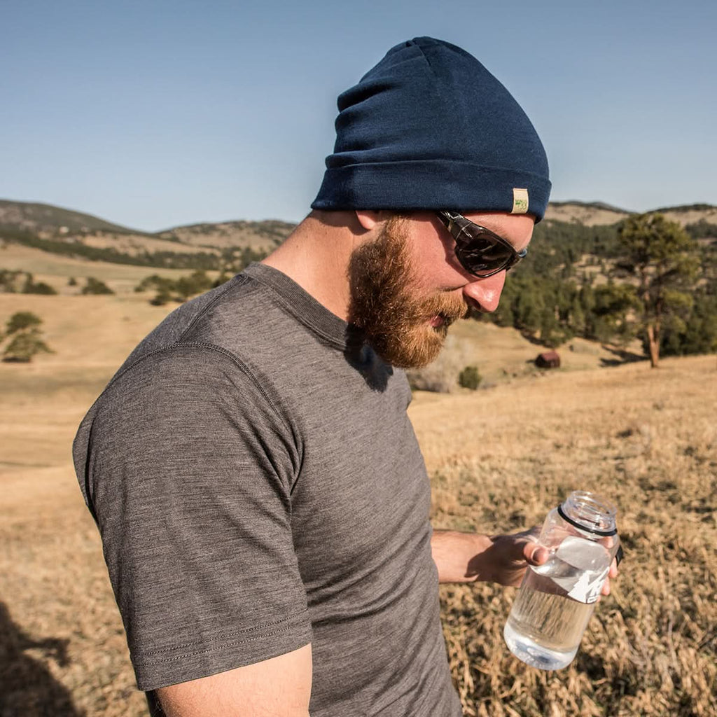 A bearded man wearing a beanie and sunglasses and a charcoal gray 100 percent merino wool t shirt holding a water bottle in an open grassland.