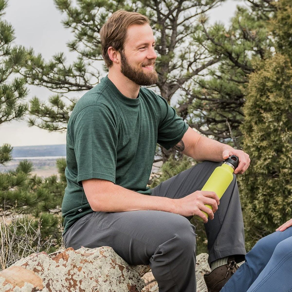  A bearded man wearing a forest green 100 percent merino wool t shirt resting on a boulder with a water bottle outdoors.