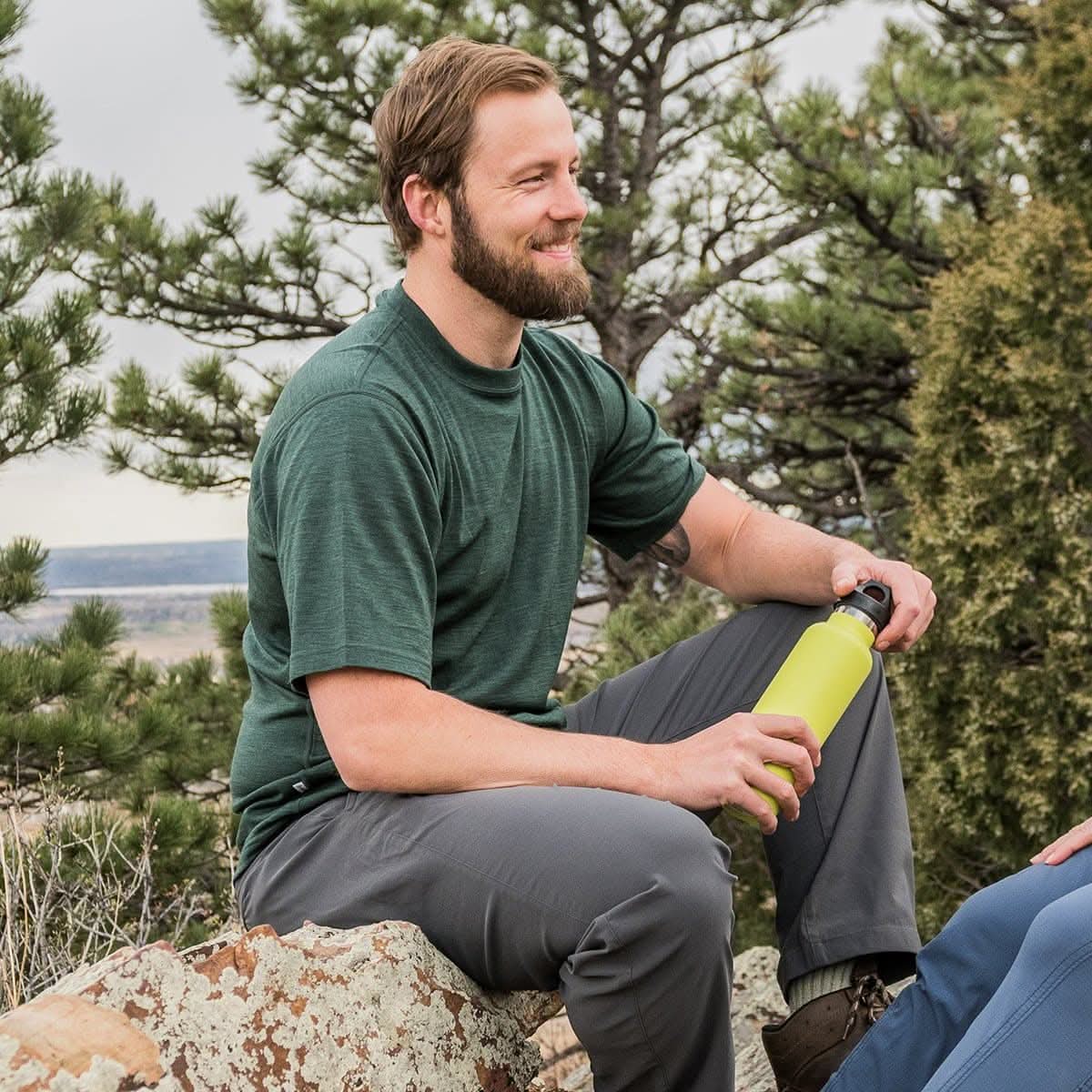  A bearded man wearing a forest green 100 percent merino wool t shirt resting on a boulder with a water bottle outdoors.