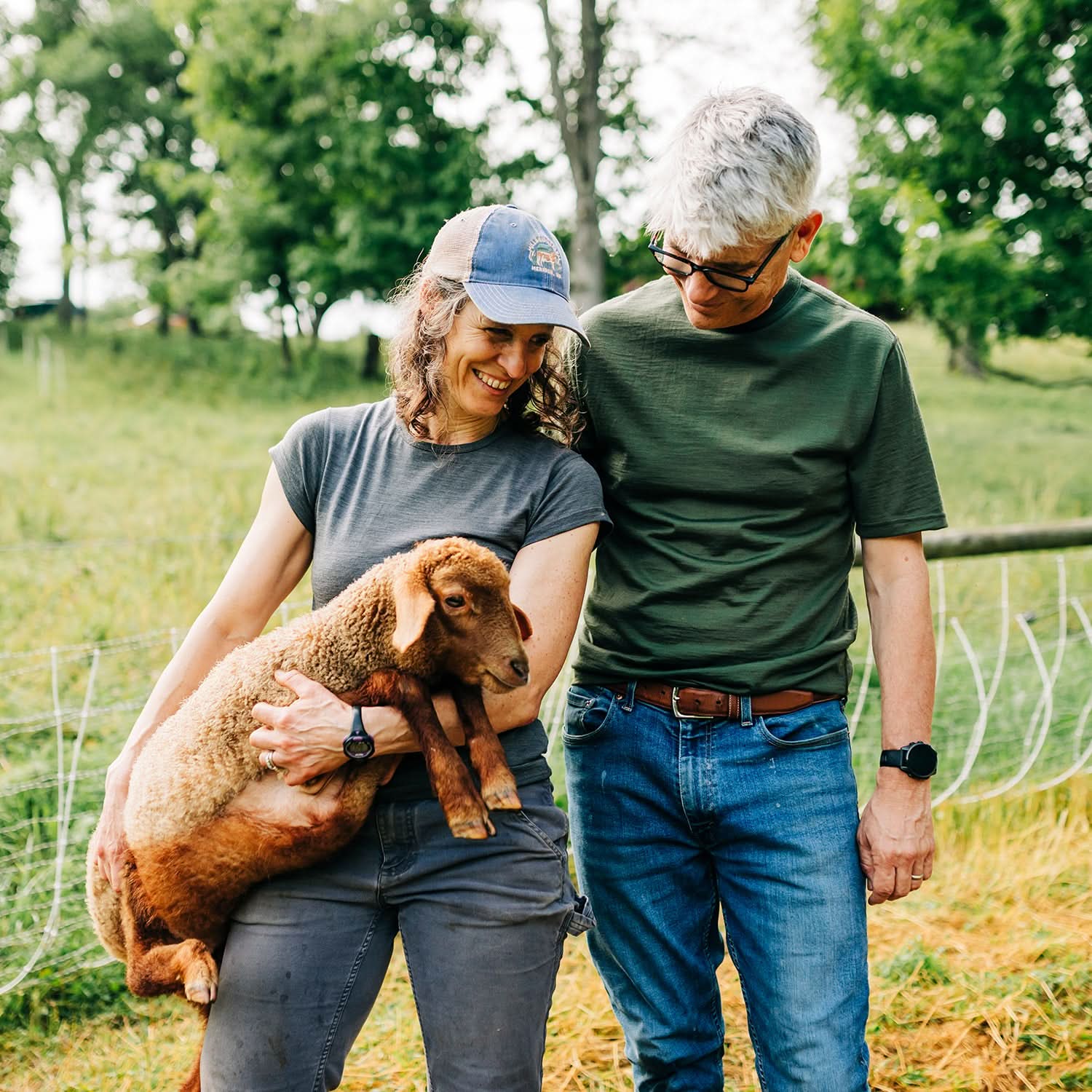 A man wearing an olive drab green 100 percent merino wool t shirt standing next to a woman holding a puppy outdoors on a farm.