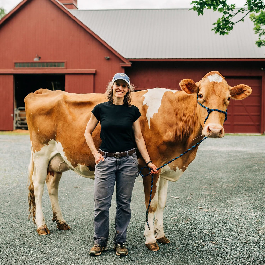 A woman wearing a cap and a black 100 percent merino wool t shirt standing next to a dairy cow outside on a farm.