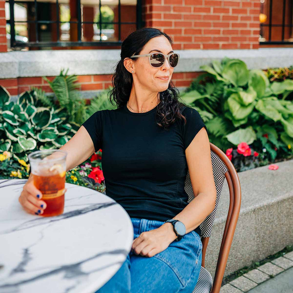 A woman wearing sunglasses and a black 100 percent merino wool t shirt sitting outdoors at a table holding an iced drink.