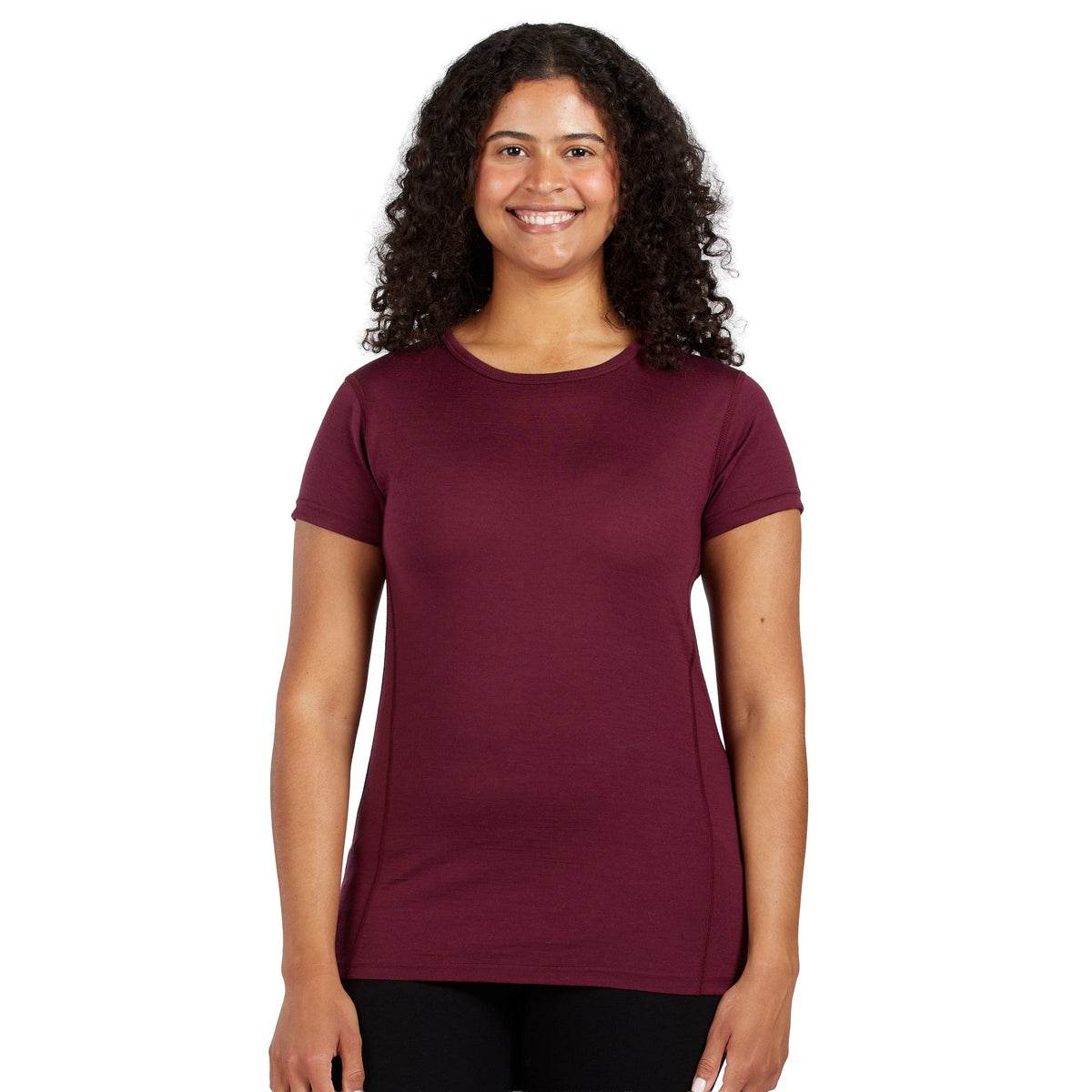A smiling woman with curly shoulder length hair wearing a burgundy 100 percent merino wool t shirt shown from the waist up against a solid white background.