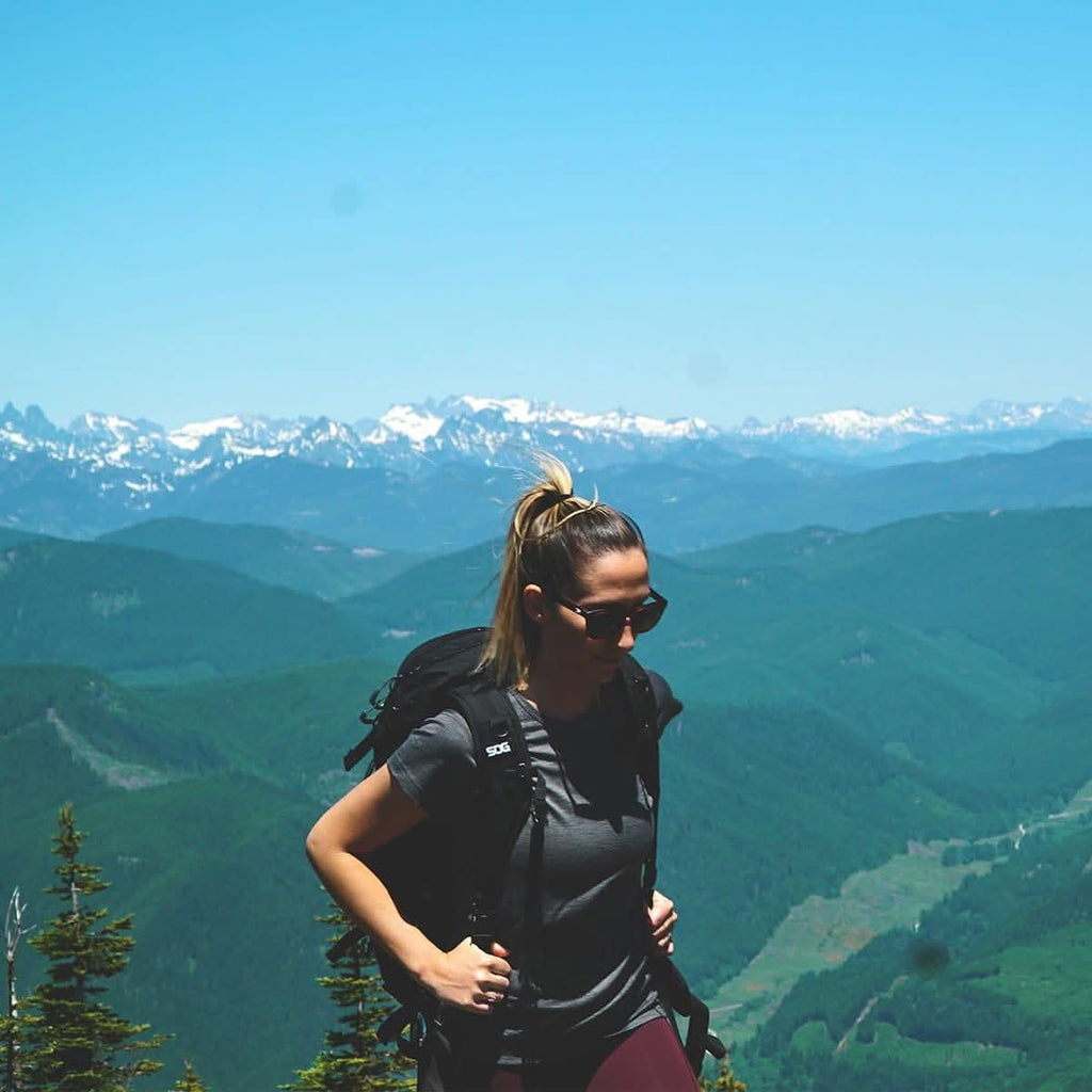 A woman wearing a 100 percent merino wool t shirt and backpack standing on a mountain summit overlooking scenic alpine peaks on a sunny day