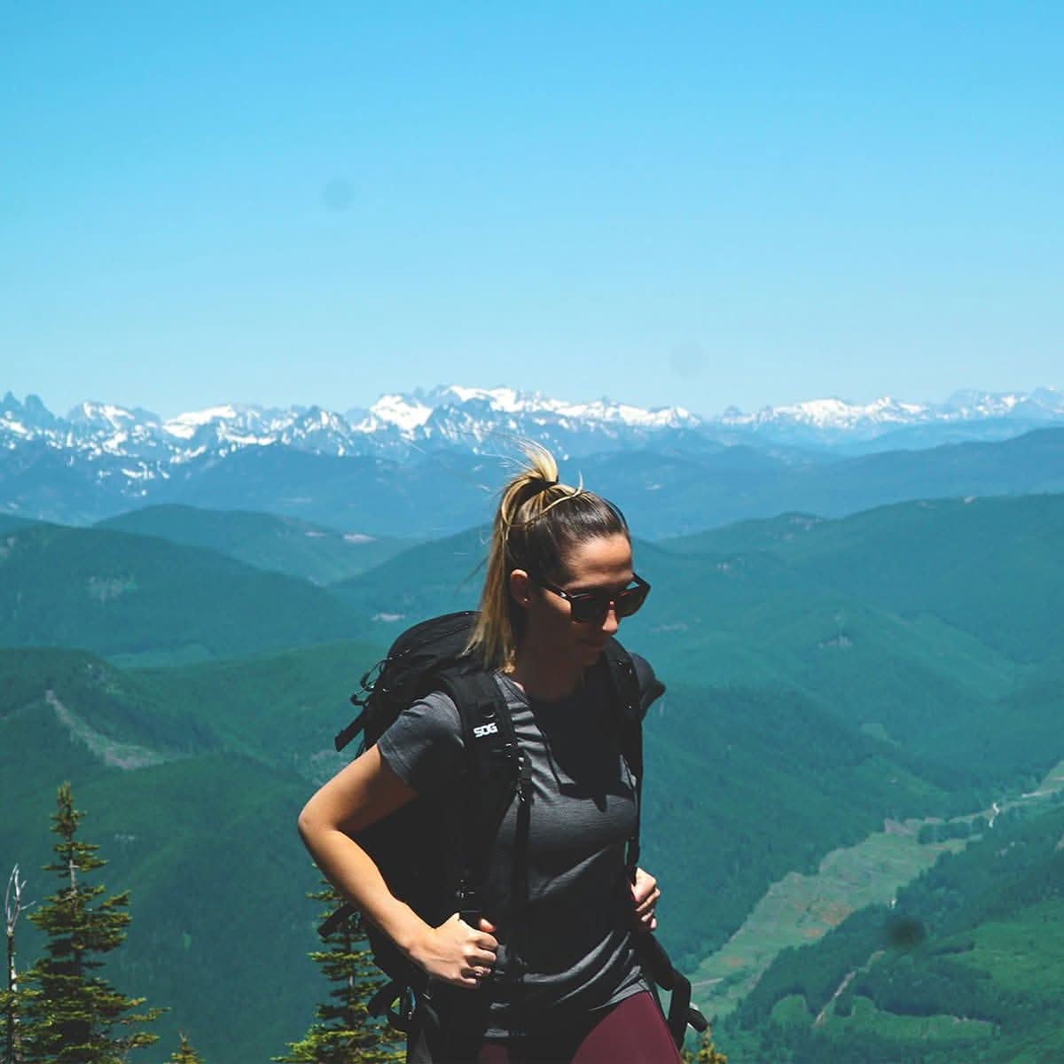 A woman wearing a 100 percent merino wool t shirt and backpack standing on a mountain summit overlooking scenic alpine peaks on a sunny day