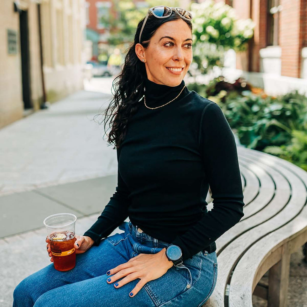 A woman wearing a black 100 percent merino wool turtleneck sitting outside on a curved bench holding an iced drink.