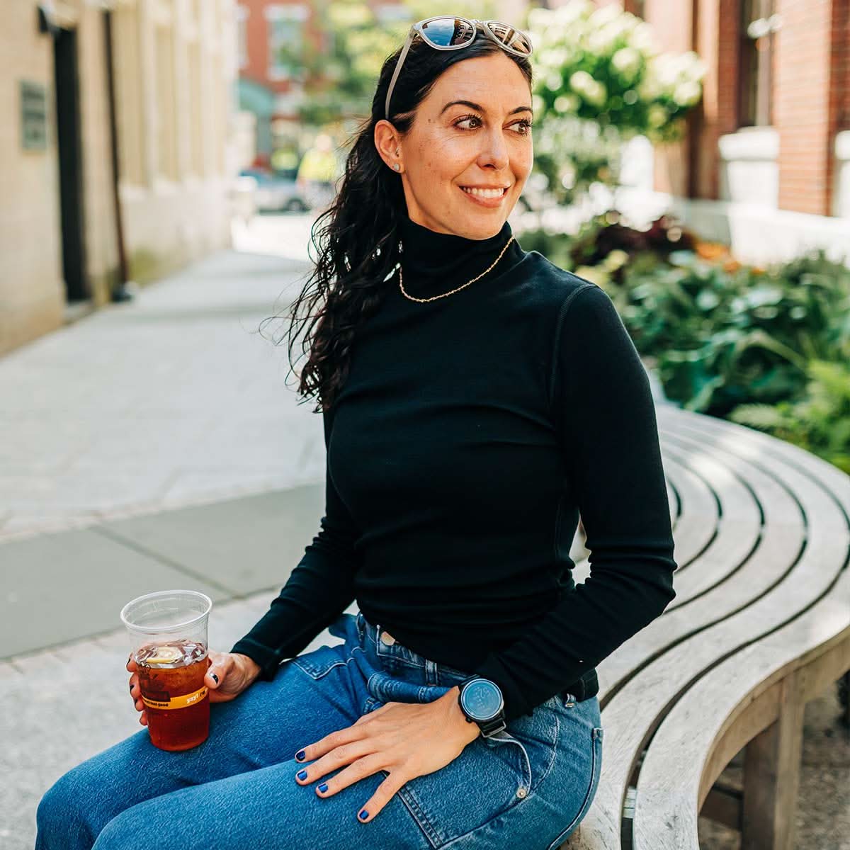 A woman wearing a black 100 percent merino wool turtleneck sitting outside on a curved bench holding an iced drink.