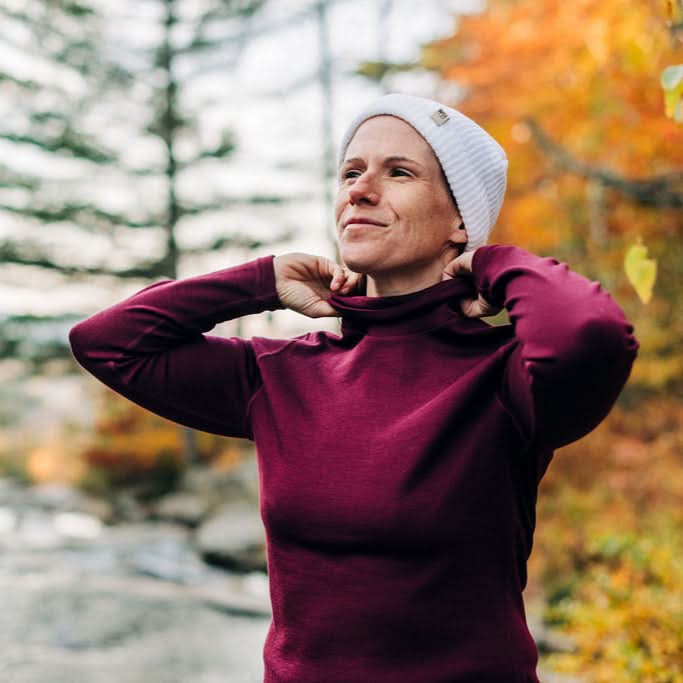 A woman wearing a burgundy 100 percent merino wool turtleneck and grey beanie standing outdoors in an autumn forest.