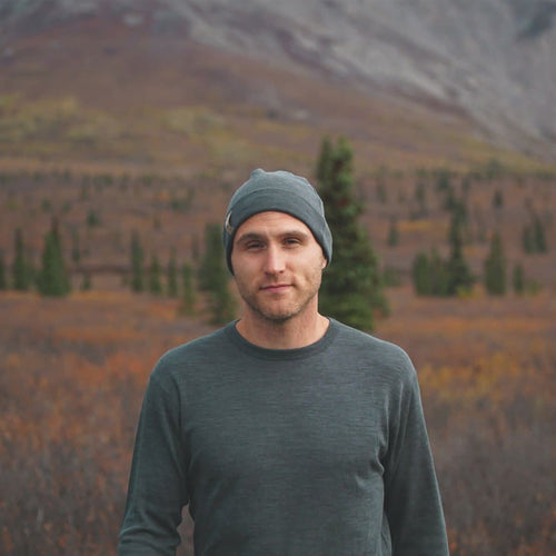 Man wearing a gray beanie and charcoal gray 100% merino wool base layer top  standing in a mountainous landscape with trees and grass.