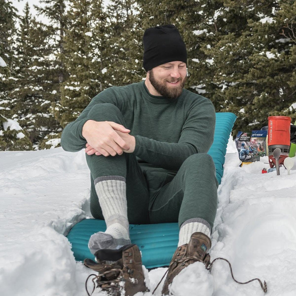 Man sitting on a blue inflatable chair in the snow with trees in the background wearing a 100% merino wool forest green base layer top