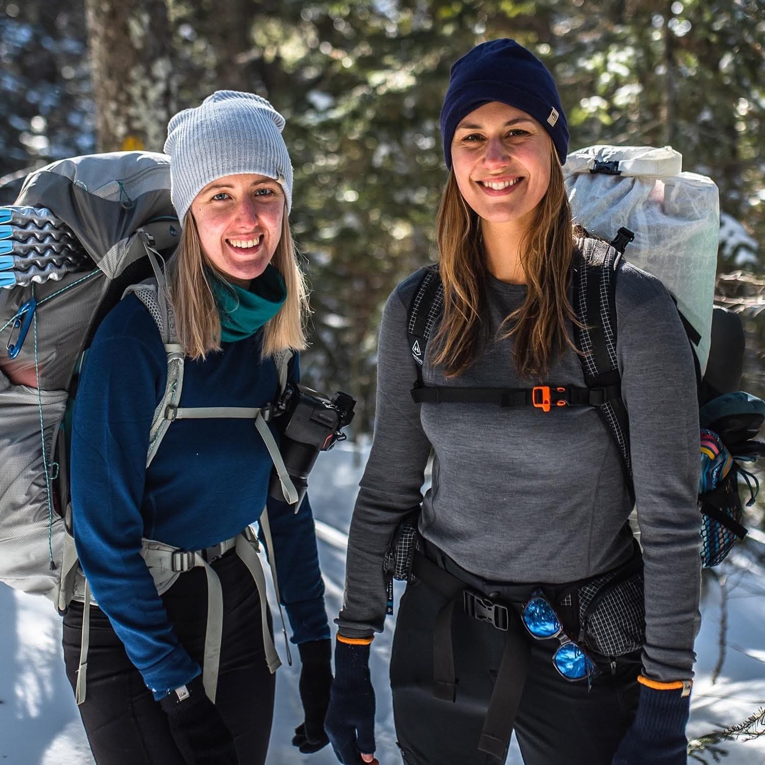 Two smilin women with backpacks in a snowy forest wear 100 percent merino wool base layer tops in blue and gray