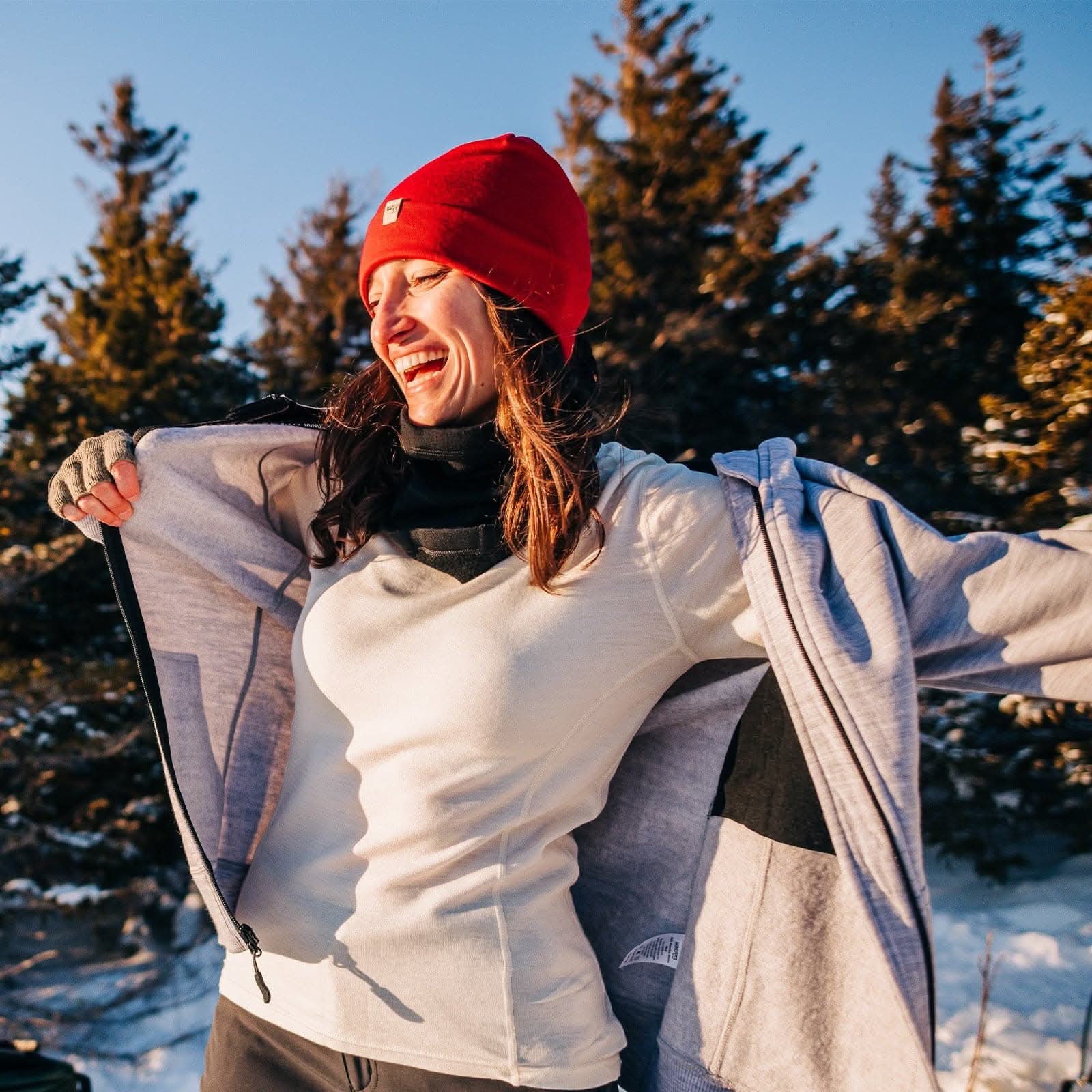 smiling woman wearing a red beanie and white 100 percent merino wool base layer top in the sun with snow and trees