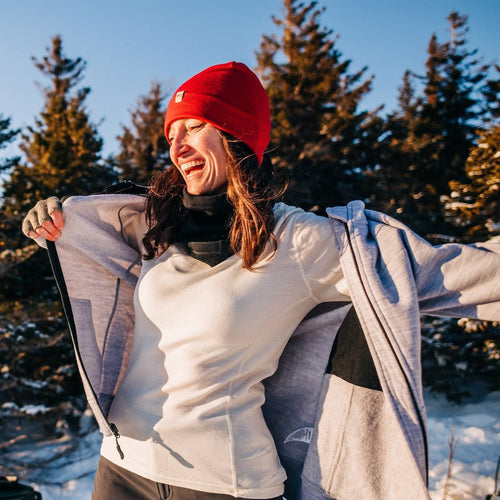 smiling woman wearing a red beanie and white 100 percent merino wool base layer top in the sun with snow and trees