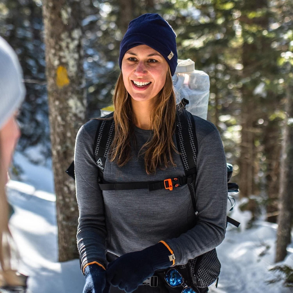 Woman in winter gear smiling in a snowy forest wearing a gray 100 percent merino wool base layer top