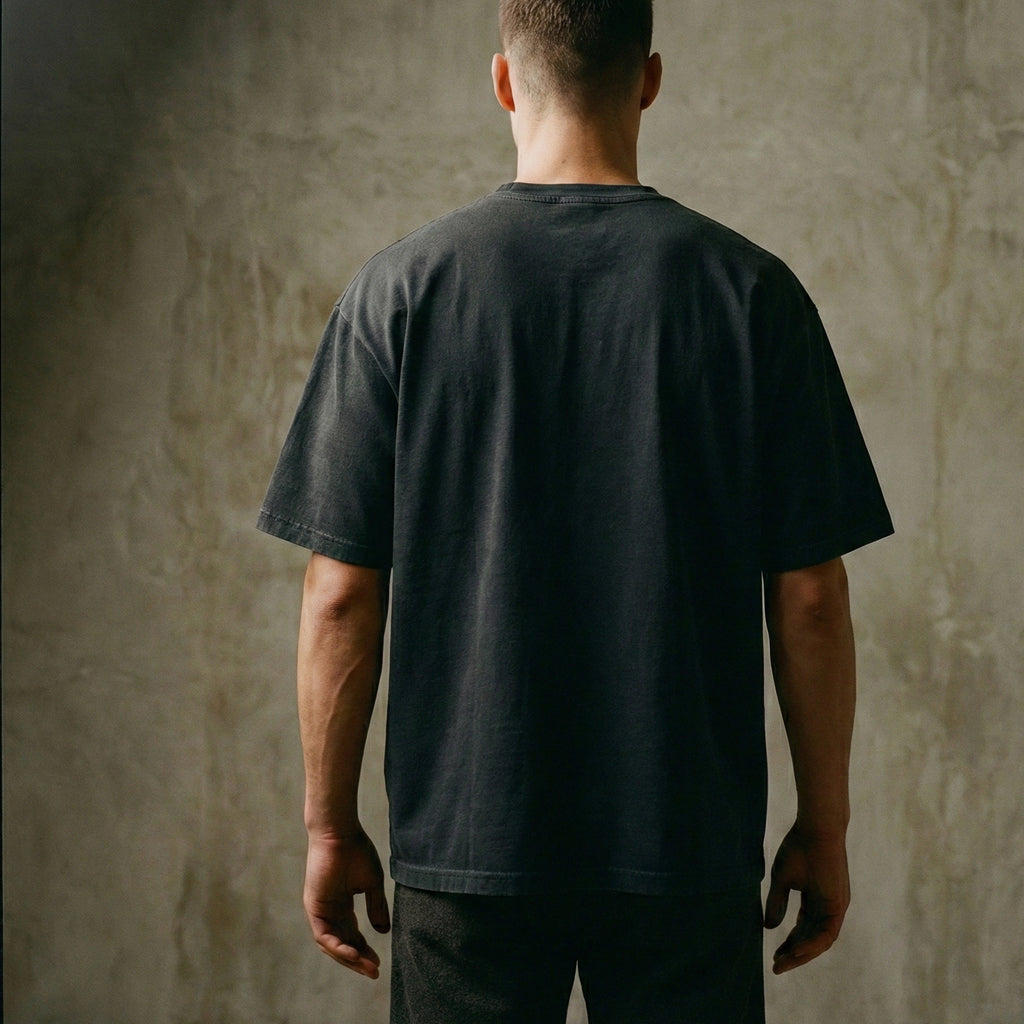 Man wearing a garment-dyed faded black oversized carded-cotton t-shirt against a textured concrete background - view from behind