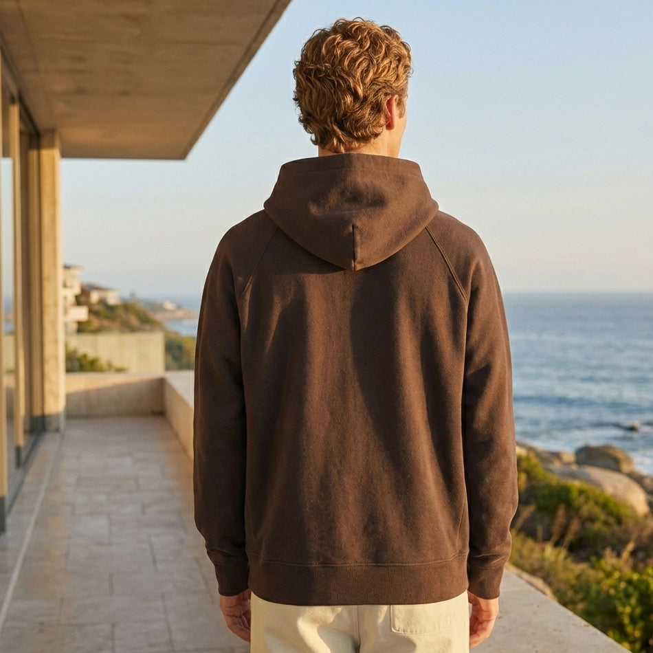Person wearing a brown hoodie standing on a balcony overlooking the ocean.